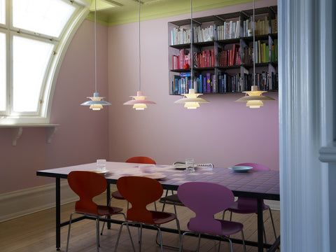 Four Louis Poulsen PH 5 Opal pendant lights in a row above a dining table with red and violet chairs, in a pink room with an arched window