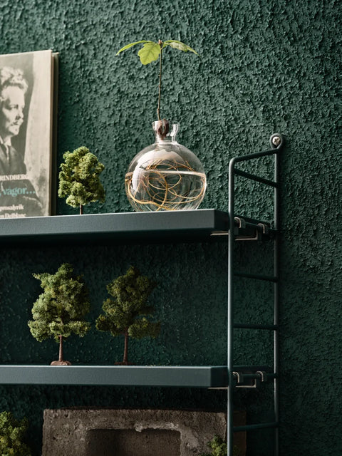 Green String® Pocket shelf styled with miniature trees and glass vase against textured green wall – Scandinavian modular shelving