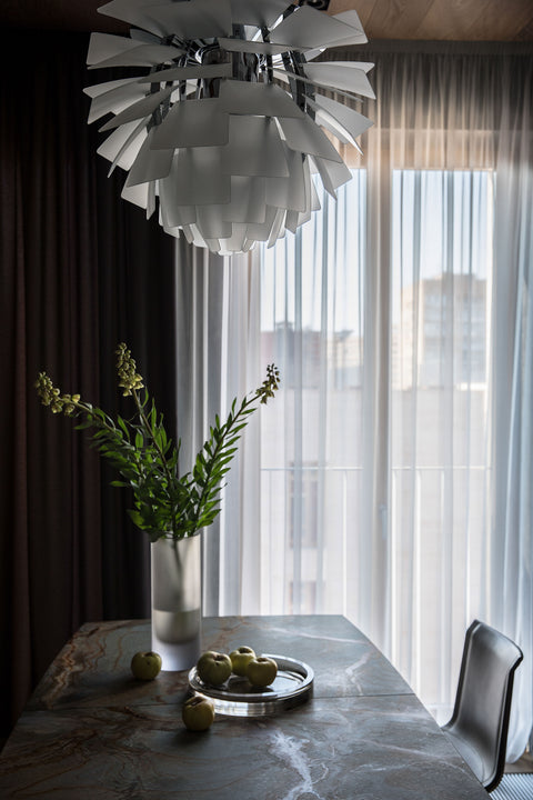 Close up of the PH Artichoke pendant over a dining table with frosted glass vase and apples against sheer window curtains