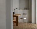 Frama Tasca Chair in Aluminum, chair with a rounded back pulled up to a round wooden table in a plaster walled room