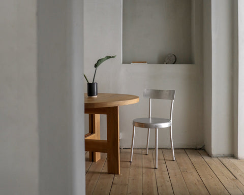 Frama Tasca Chair in Aluminum, chair with a rounded back pulled up to a round wooden table in a plaster walled room