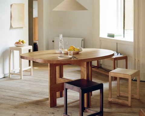 Frama Low Stool 01 in Natural Birch, pale birch stools and one dark stool set around an oval wooden table under a pendant shade