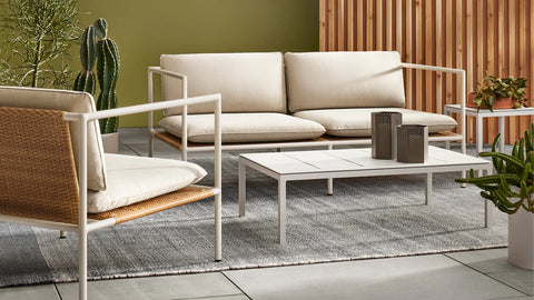 Blu Dot Dog Days Outdoor 2 Seat Sofa in Sunbrella Linen cushions with white powder-coated aluminum frame and all-weather rattan, styled on a patio with matching lounge chair, coffee table, and cacti