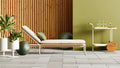 Blu Dot Dog Days Outdoor Sun Lounger in Sunbrella Linen cushion with white powder-coated aluminum frame and all-weather rattan, styled on a patio with cedar wood wall, green potted plants, and matching bar cart