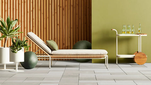Blu Dot Dog Days Outdoor Sun Lounger in Sunbrella Linen cushion with white powder-coated aluminum frame and all-weather rattan, styled on a patio with cedar wood wall, green potted plants, and matching bar cart