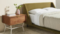 Blu Dot Dang 1 Drawer Nightstand in walnut veneer with putty powder-coated steel legs and antique brass drawer pull, styled in a modern bedroom beside a moss green upholstered bed with a pink vase and stacked books on top