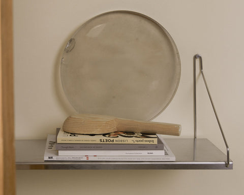 Frama Single Shelf, Stainless Steel, shelf holding a stack of books and a wooden paddle with a round metal tray leaning behind