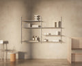 Frama Shelf Library Double Section in Stainless Steel, steel shelving on a plaster wall holding folded towels, bottles and cups, chair to one side