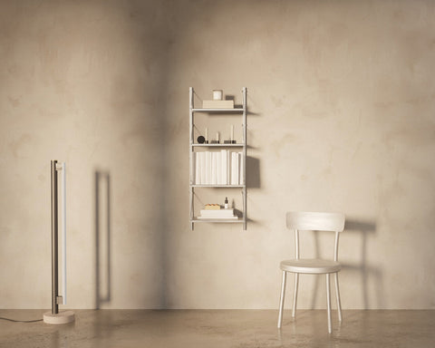 Frama Shelf Library Add-on Section in Stainless Steel, narrow steel shelving on a plaster wall holding books and boxes, with a wooden chair nearby