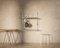 Frama Shelf Library Hanger Section in Stainless Steel, steel shelves holding bottles with a pan and utensils hanging, trestle table and stool nearby