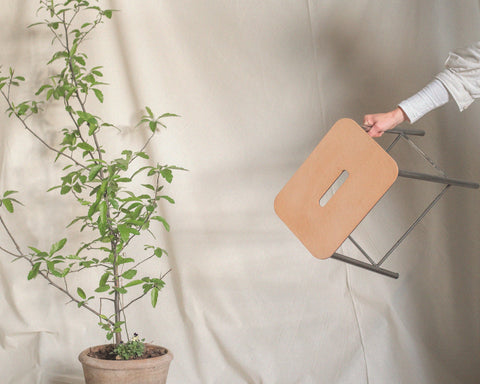 Frama Adam Stool, a hand lifting the leather seat top by its slot handle in front of a draped cloth and a potted plant
