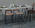 Frama Adam Stool, four stools pulled up to a long table set with candles and cups, a bentwood chair at the far end