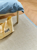 HAY Shopper Bag, Soft Yellow, cotton tote bag, pale yellow shopper on a cream rug beside a bed with blue striped bedding
