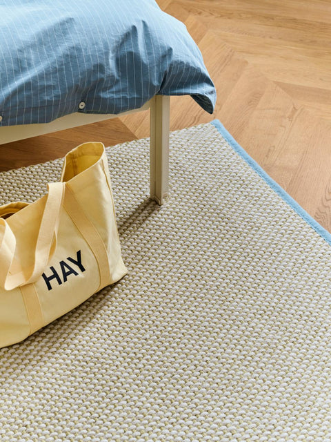 HAY Shopper Bag, Soft Yellow, cotton tote bag, pale yellow shopper on a cream rug beside a bed with blue striped bedding