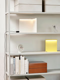 HAY Table Clock, White, white table clock, on a white shelving unit beside a yellow glowing lamp, books and a tan box