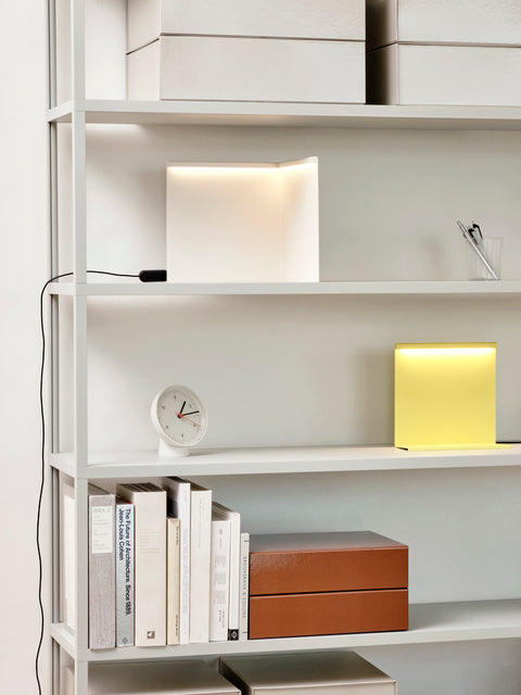 HAY Table Clock, White, white table clock, on a white shelving unit beside a yellow glowing lamp, books and a tan box
