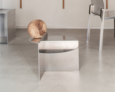 Frama Rivet Side Table in Aluminum, metal table on a concrete floor beside a round wooden seat with a chair leg in frame