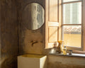 Frama CM-1 Circle Mirror, round mirror on a worn plaster wall above a pale stone basin beside a shuttered window