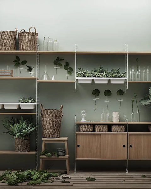 String Furniture cabinet with sliding doors in oak, part of a Scandinavian shelving system styled with plants and storage baskets