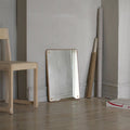 Frama RM-1 Rectangular Mirror, mirror leaning against a grey wall on a plank floor beside a pale wood chair and paper rolls