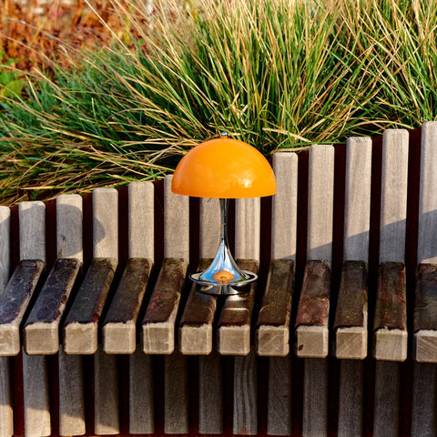 Louis Poulsen Panthella 160 portable lamp in orange displayed on a wooden bench with greenery behind