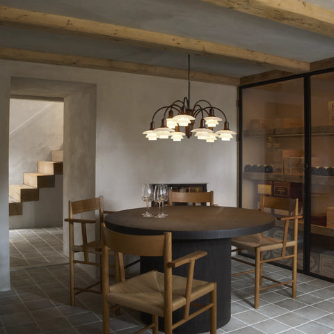 Louis Poulsen PH 1/1 Chandelier hanging above a dark round pedestal dining table with rush-seat armchairs in a stone-walled room with exposed timber beams and glass wine cellar