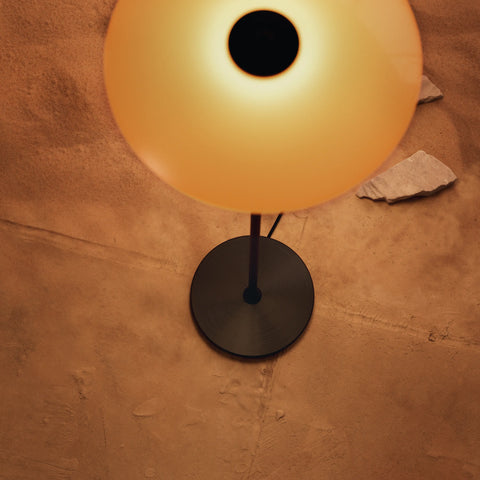 PH 3½-2½ Floor Lamp Centenary Edition — amber glass upper shade glowing warmly, showing dark circular base on textured wall background