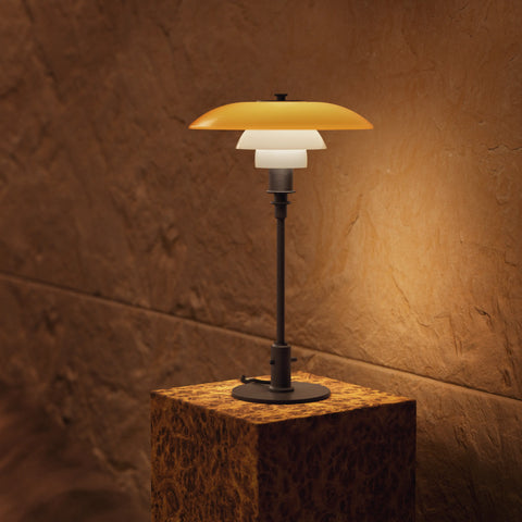 Louis Poulsen PH 3/2 Table Lamp Centenary Edition in aged brass with amber and opal glass shades, illuminated on a stone pedestal against a warm terracotta wall