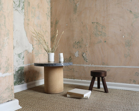 Frama AML Stool in Dark Pine, dark stool on a sisal rug beside a stone pedestal table and dried grasses in a stripped room