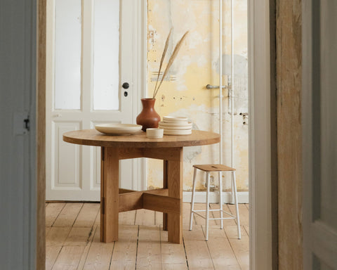 Frama Farmhouse Trestle Table in Natural Oak, round oak table with a vase and stacked bowls seen through a doorway, stool alongside