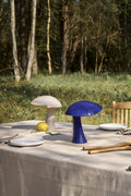 Rumee 220 Portable Lamps in Earth Grey and Night Blue placed on an outdoor dining table in a forest setting.