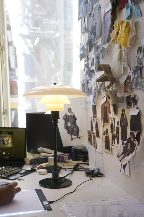 Louis Poulsen PH 2/1 Table Lamp in Glossy Deep Green, lit on a designer's desk beside a wall of pinned fabric swatches and sketches