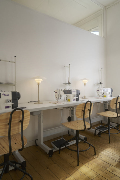 Two Louis Poulsen PH 3/2 Glass Table Lamps in Glossy Beige lit along a sewing atelier worktable with machines, wooden chairs and a plank floor