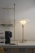 Louis Poulsen PH 3/2 Glass Table Lamp in Glossy Beige, lit on a workbench beside an industrial sewing machine
