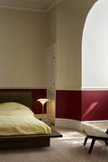 Louis Poulsen PH 3/2 Glass Table Lamp in Glossy Beige lit at a bedside, seen through a curved archway into a bedroom with a deep red painted dado