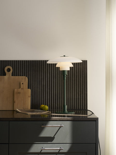 Louis Poulsen PH 3/2 Glass Table Lamp in Glossy Deep Green, lit on a dark kitchen counter beside wooden chopping boards and lemons