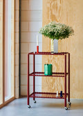 HAY Arcs Vase, Small, Green, green metal flower vase with scalloped sides, on a deep red trolley below a mirror vase of white blossom
