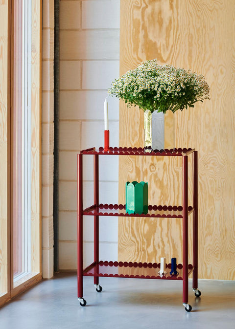 HAY Arcs Vase, Small, Green, green metal flower vase with scalloped sides, on a deep red trolley below a mirror vase of white blossom