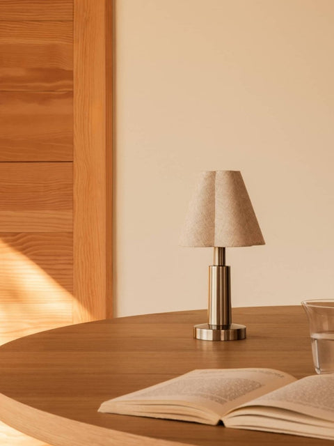 Audo Copenhagen Admiral Portable Lamp with a sculpted fabric shade and metallic base sits on a wooden dining table beside an open book