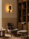 Bronzed aluminium Wing Wall Lamp lit above a lounge chair and ottoman beside an oak bookshelf