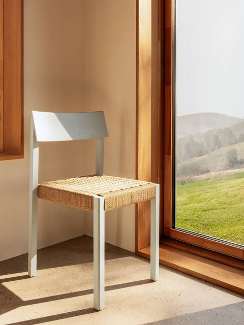 Audo Copenhagen Mauro Dining Chair in Linen Blue Oak with a woven paper cord seat, standing in a sunlit corner by a window