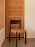 Audo Copenhagen Mauro Dining Chair in French red oak with a handwoven paper cord seat, set against a minimalist wooden door