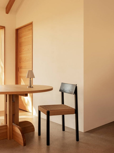 Audo Copenhagen Mauro Dining Chair, black oak Mauro Dining Chair with a paper cord seat beside a Puffin Dining Table and Admiral Portable Table Lamp