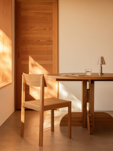 Audo Copenhagen Admiral Portable Lamp on a wooden Puffin Dining Table beside a Mauro Dining Chair in a warm, sunlit room