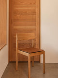 Audo Copenhagen Mauro Dining Chair in natural oak with a brown leather seat cushion, positioned against a wooden door and light wall
