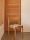 Audo Copenhagen Mauro Dining Chair Seat Cushion in light textile on a wooden Mauro Dining Chair against a warm timber door