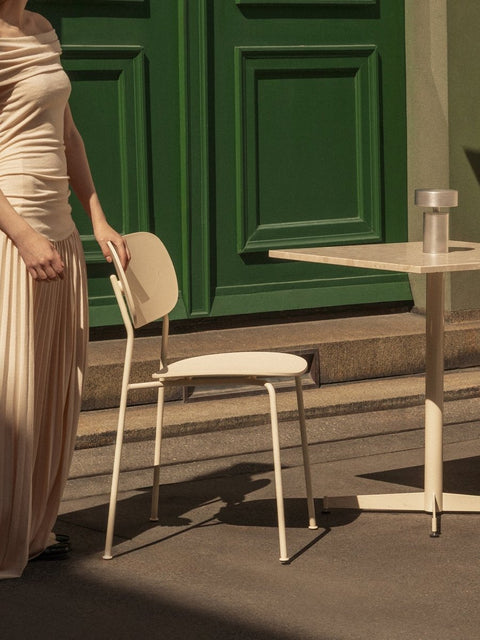 Audo Copenhagen Co Dining Chair, Outdoor, person in a beige dress by an ivory Co Dining Chair, Outdoor, Nook Dining Table and Column Portable Lamp by a green door