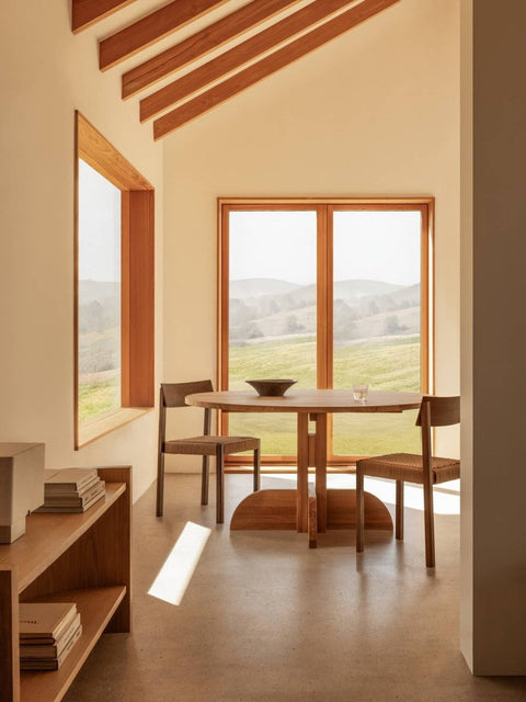 Audo Copenhagen Puffin Dining Table, Round, oak Puffin Dining Table and Mauro Dining Chairs in a sunlit room with Crescent Shelving and views of rolling green hills
