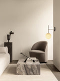Audo Copenhagen Tearoom Swivel Chair, With Loose Cover, grey Tearoom Lounge Chair sits by a Plinth Low marble table. A TR Bulb wall lamp glows above, in a modern living room