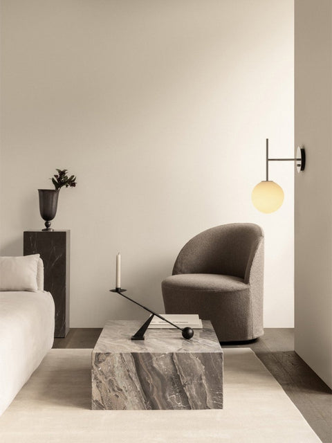 Audo Copenhagen Tearoom Swivel Chair, With Loose Cover, grey Tearoom Lounge Chair sits by a Plinth Low marble table. A TR Bulb wall lamp glows above, in a modern living room